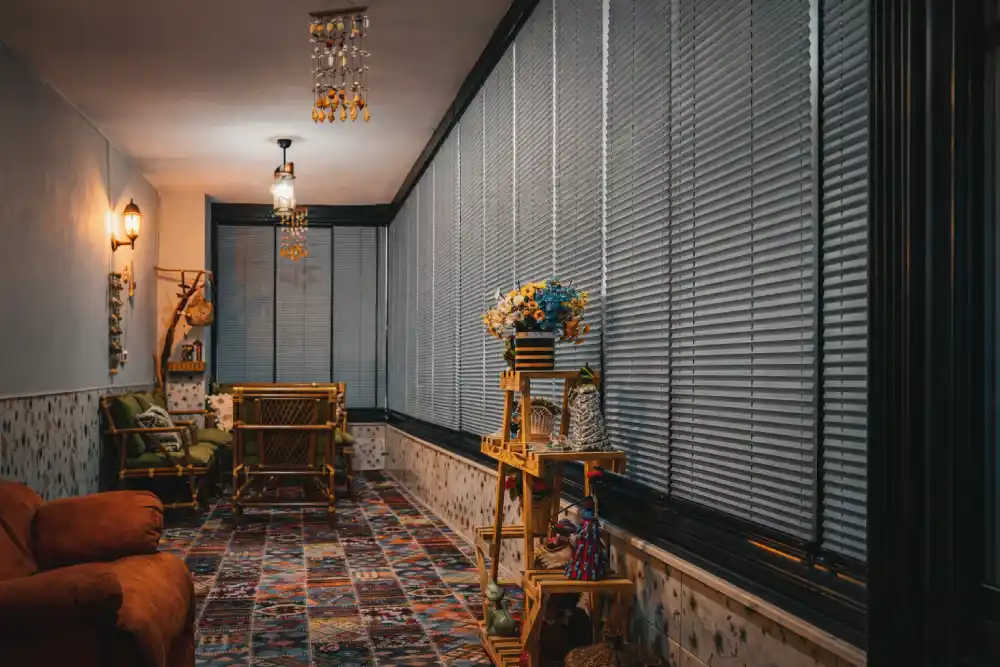 Bambu mobilyalı, desenli halılı ve tam boy yatay jaluzili kapalı balkon iç mekânı.